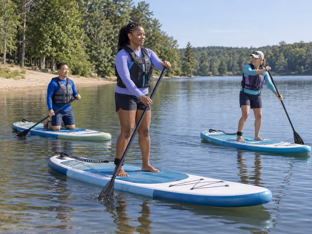 Three adults wearing flotation vests practise paddleboarding on calm water near a sandy shore.
