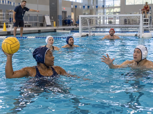 Adults in water polo caps practice passing a yellow ball in an indoor pool, with a rectangular goal, coach and lifeguard behind them.