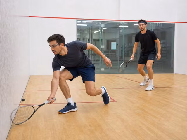 Two squash players wearing protective eyewear, with one reaching for a ball by the side wall and the other waiting clear behind him