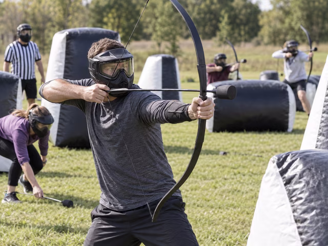 Eight adult players wearing fitted full-face masks use purpose-made bows and broad cylindrical foam-tipped arrows behind padded barriers on a professionally supervised divided field.