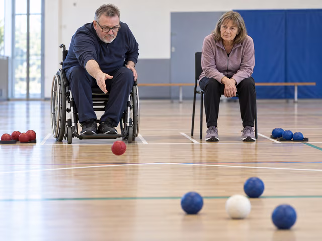 Two seated adults, one in a wheelchair and one on a stable chair, carefully place soft red and blue boccia balls toward a white jack on a taped indoor boccia court.