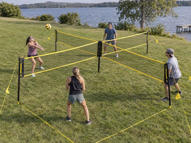 Four adults each occupy a different square around a cross-shaped volleyball net on level grass, with one ball in the air and visible support-line markers.