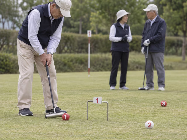 Adult teammates on a marked short-grass gateball court use T-headed mallets with numbered red and white balls, three small gates and a center pole; one prepares a careful shot while teammates plan.
