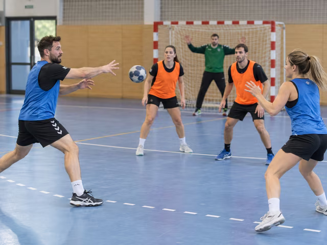 Adult handball players in two bib colors pass a small ball outside a clearly marked goal-area arc, with a goalkeeper in an anchored indoor handball goal.
