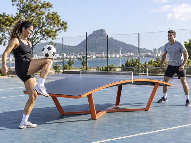 Two adults use a thigh and foot to rally a football across a curved TEQ table in a clear recreation court; no paddles and no hands touching the live ball.